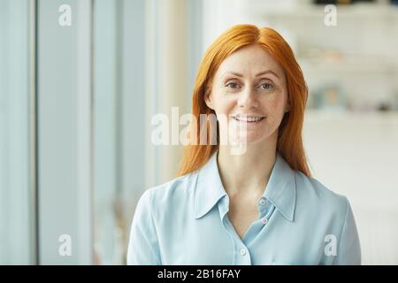 Kopf- und Schulterporträt der rothaarigen Geschäftsfrau lächelt an der Kamera, während sie im modernen weißen Büro am Fenster steht, Kopienraum Stockfoto