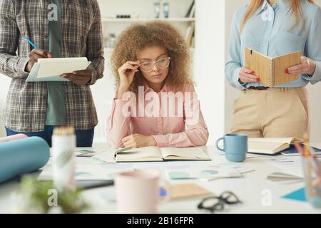 Portrait einer intelligenten jungen Frau, die große Brille trägt und Notizen im Planer liest, während sie mit einem kreativen Team an einem Geschäftsprojekt arbeitet Stockfoto