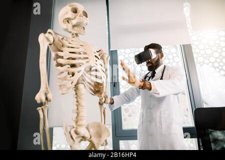 Ein junger afrikanischer Arzt, Wissenschaftler oder Medizinstudent verwendet eine erweiterte Realitätsbrille, um das menschliche Skelett zu eruden. Virtuelle Realität, Anatomie, Medizin Stockfoto