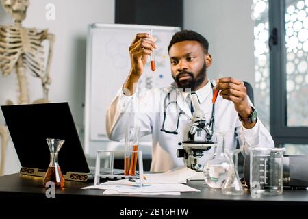 afroamerikanischer Wissenschaftler mikrobiologischer Chemiker im weißen Mantel, der Reagenzgläser mit Reagenzien hält und untersucht, sitzt am Tisch mit Laptop, m Stockfoto