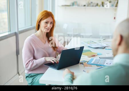 Portrait einer reifen, rothaarigen Geschäftsfrau, die einen Mann interviewt, während sie einen Laptop im Büro verwendet, Platz für Kopien Stockfoto