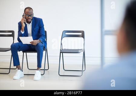 Komplettes Portrait eines modernen afroamerikanischen Mannes, der auf ein Bewerbungsgespräch hinter der Glaswand wartet und per Telefon spricht, Copy-Space Stockfoto