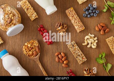 Draufsicht über die Lebensmittelzusammensetzung von Beeren, Nüssen, Müsli und Cornflakes sowie Flaschen Joghurt und Milch auf Holzgrund Stockfoto