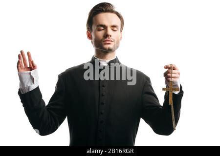 Konzentrierter katholischer Priester, der mit erhobener Hand betet und dabei goldenes Kreuz isoliert auf Weiß hält Stockfoto