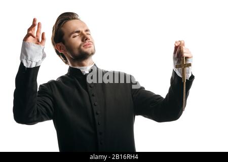 Junger katholischer Priester, der mit geschlossenen Augen und erhobenen Händen betete und dabei goldenes Kreuz auf weißem hielt Stockfoto