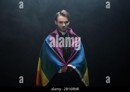 Der seriöse katholische Priester ist in die lgbt-Flagge gehüllt, während er die Kamera auf schwarzem Hintergrund mit Rauch betrachtet Stockfoto