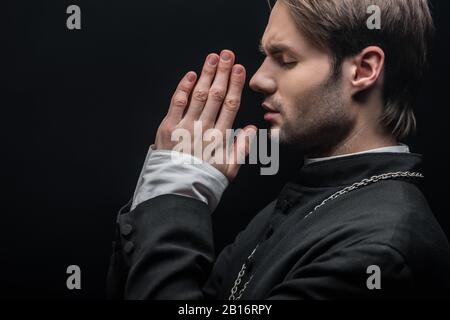 Seitenansicht des jungen konzentrierten katholischen Priesters, der mit geschlossenen Augen auf Schwarz bettete Stockfoto