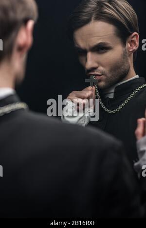 Der junge, angespannte katholische Priester küsst das heilige Kreuz, während er seine eigene Reflexion betrachtet, die auf Schwarz isoliert ist Stockfoto