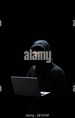 Junger Hacker in der Haube, der Laptop isoliert auf Schwarz verwendet Stockfoto