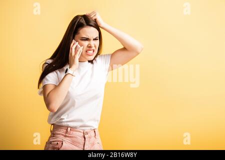 Irritierte Mädchen berühren Kopf und schreien, während sie auf dem Smartphone sprechen, isoliert auf Gelb Stockfoto