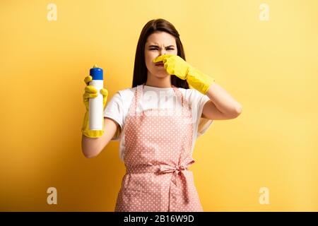 Unzufriedener Hausmann in der Schürze und Gummihandschuhe verstopfen den Schlauch mit der Hand, während der Lufterfrischer auf gelbem Hintergrund gehalten wird Stockfoto