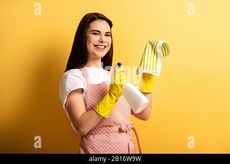 Fröhliche Hausfrau in der Schürze und Gummihandschuhe, die Sprühflasche und Lappen auf gelbem Hintergrund halten Stockfoto