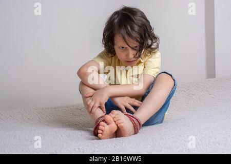 Kleiner Junge mit Füßen, die mit Seil in emotionalem Stress zusammengebunden sind Stockfoto