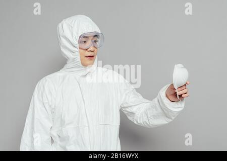 Asiatischer Epidemiologe im Hazmatanzug, der Atemschutzmaske auf grauem Hintergrund hält Stockfoto