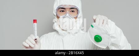 Panoramaaufnahme des asiatischen Epidemiologen mit Atemschutzmaske und Reagenzglas mit auf Grau isolierter Blutprobe Stockfoto