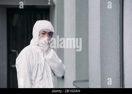 Asiatischer Epidemiologe im Hazmatanzug und in der Atemschutzmaske, der die Kamera betrachtet, während er draußen steht Stockfoto