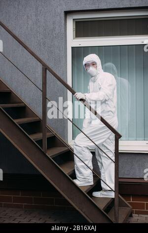 Asiatischer Epidemiologe im Hazmatanzug und Atemschutzmaske, der nach oben vor dem Gebäude spaziert Stockfoto