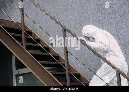 Asiatischer Epidemiologe im Hazmatanzug und Atemschutzmaske, der nach oben vor dem Gebäude spaziert Stockfoto