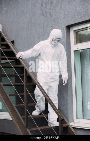 Asiatischer Epidemiologe im Hasmatanzug und Atemschutzmaske, der unten vor dem Gebäude spazieren ging Stockfoto