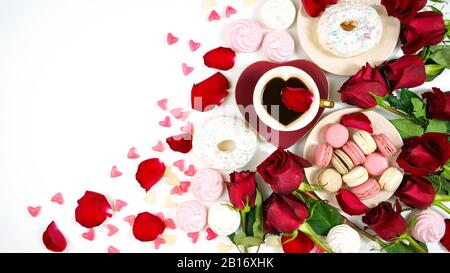 Rote Rosen und Morgentee kreative Komposition flaches Lay-Layout mit Kaffee in herzförmiger Tasse und Untertasse sowie Makronen und Donuts auf weißem Hintergrund, Stockfoto