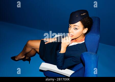 Hochwinkeliger Blick auf den afroamerikanischen Flugbegleiter, der auf dem Sitz auf blauem Hintergrund sitzt Stockfoto