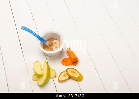 Hochwinkeliger Blick auf Babynahrung in Schüssel mit Löffel und geschnittenem Obst und Karotten auf weißer Holzoberfläche Stockfoto