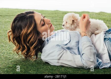 Frau, die auf Gras liegt und Havanese Welpen betrachtet, isoliert auf Weiß Stockfoto