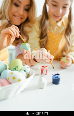 Selektive Fokussierung von dekorativen Häschen in der Nähe von Kind und Mutter, die Pinsel beim Malen von ostereiern halten Stockfoto