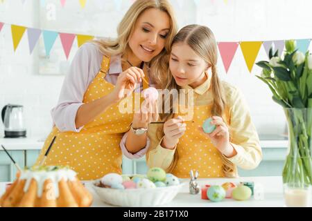 Selektiver Fokus von Mutter und Tochter beim Malen von Hühnereiern in der Nähe gebackener osterkuchen, Tulpen und dekorativem Kaninchen Stockfoto