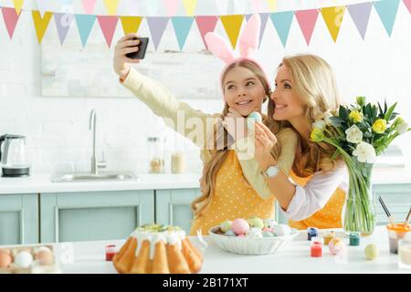 Glückliche Mutter und Tochter in Hünenohren, die selfie in der Nähe von osterkuchen einnehmen und Hühnereier bemalten Stockfoto