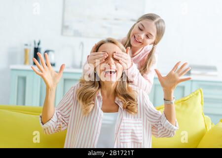 Glückliches Kind, das die Augen der überraschten Mutter im Wohnzimmer bedeckt Stockfoto