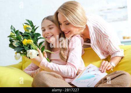 Glückliche Mutter hält Tulpen und Grußkarte mit 8. märz Schriftzug in der Nähe lächelnder Tochter zu Hause Stockfoto