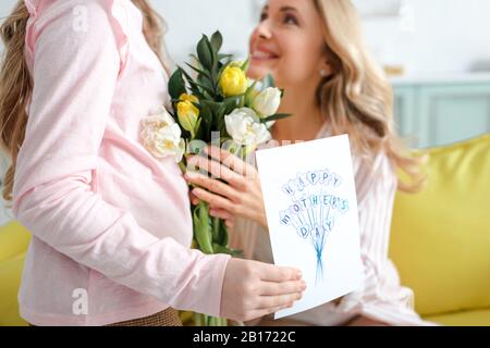 Selektive Fokussierung des Kindes mit Grußkarte mit Happy Mothers Day Schriftzug und Blumen in der Nähe einer glücklichen Mutter Stockfoto