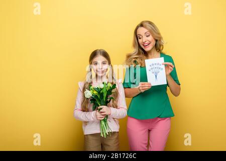 Glückliche Mutter hält Grußkarte mit Happy Mothers Day Schriftzug in der Nähe niedlicher Tochter und Blumen isoliert auf Gelb Stockfoto