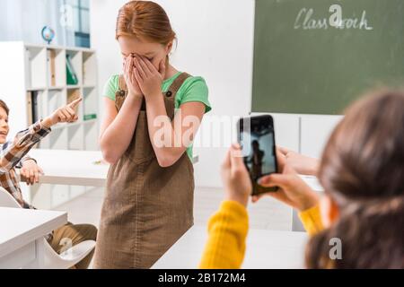 Selektive Fokussierung des Schuljungen, der mit dem Finger auf das aufgerollte Kind zeigt, während das Schulmädchen ein Foto, ein Cybermobbing-Konzept aufstellt Stockfoto