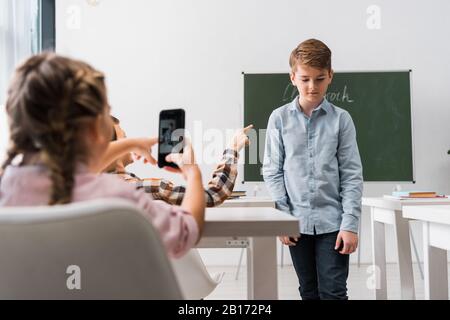Selektive Fokussierung des Schuljungen, der mit dem Finger auf den Mitschüler zeigt, während das Schulmädchen ein Foto, ein Cybermobbbingkonzept aufstellt Stockfoto