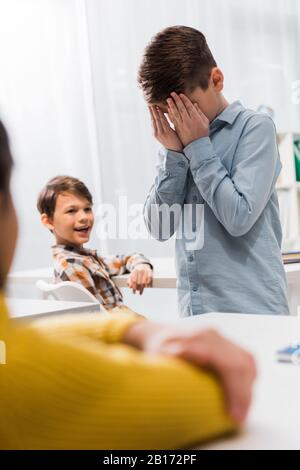 Selektive Fokussierung von glücklichen Schulkindern auf aufgebrachten Schuljungen, der Gesicht bedeckt, Mobbingkonzept Stockfoto
