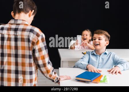 Selektive Fokussierung des Schulmädchens, das ein Foto von Klassenkameraden aufgenommen hat, während der Schuljunge isoliert über das schwarze, Cyberbullying-Konzept lachte Stockfoto