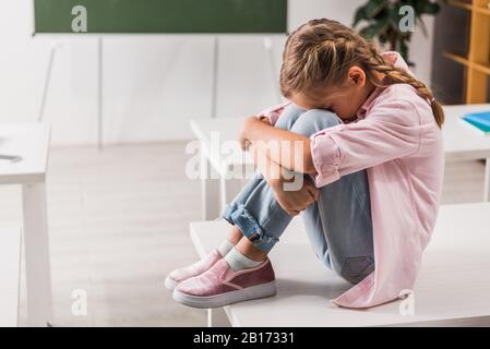 Schikanierte Schulkinder, die das Gesicht bedecken, während sie auf dem Schreibtisch im Klassenzimmer sitzen Stockfoto