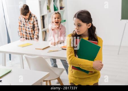 Selektive Fokussierung von schikanierten Schulmädchen, die Notizbücher in der Nähe grausamer Mitschüler halten, die mit dem Finger zeigen Stockfoto