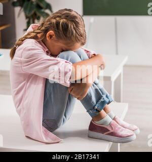 Schikanierte Schulmädchen, die Gesicht bedecken, während sie auf dem Schreibtisch im Klassenzimmer sitzen Stockfoto