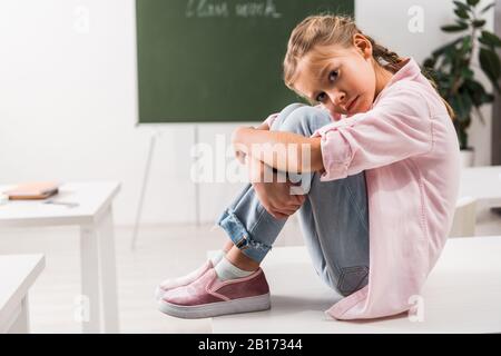 Schikanierte Schulmädchen, die auf dem Schreibtisch sitzen und die Kamera im Klassenzimmer betrachten Stockfoto