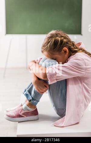 Verärgert und drangsaliert Schulmädchen, die im Klassenzimmer auf dem Schreibtisch sitzen Stockfoto