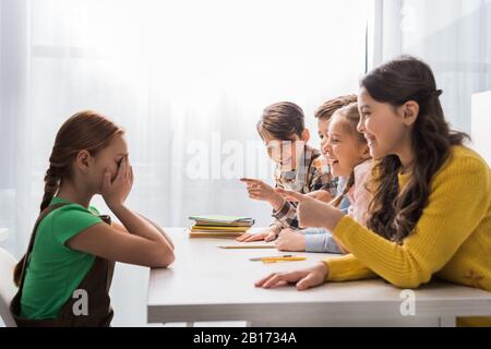 Grausame Schulkinder, die mit den Fingern zeigen und in der Nähe schikanierten Schulmädchens lachen, die Gesicht bedecken, während sie im Klassenzimmer weinten Stockfoto