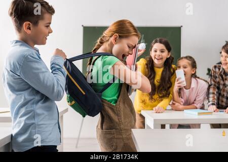 Selektive Fokussierung des Schuljungen, der den Rucksack von schikanierten Schulmädchen in der Nähe grausamer Klassenkameraden berührt, Cybermobbing Konzept Stockfoto