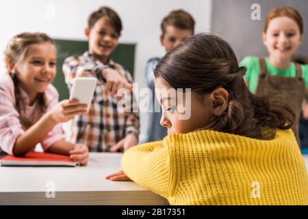 Selektive Fokussierung von schikanierten Schulmädchen in der Nähe von Schulkinds mit Smartphone und grausamen Klassenkameraden, Cyberbullying Concept Stockfoto
