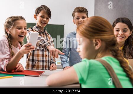 Selektive Fokussierung von Schulkinds mit Smartphone und grausamen Klassenkameraden, die in der Nähe von Schikanen lachen, Cyberbullying Concept Stockfoto