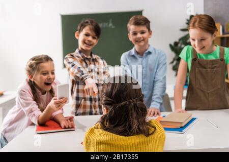 Rückansicht des schikanierten Schulmädchens, das in der Nähe eines Schulkindes sitzt, mit Smartphone und grausamen Klassenkameraden, Cybermobbing Konzept Stockfoto