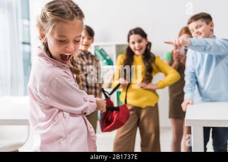 Selektive Fokussierung von schikanierten Schulmädchen, die in der Nähe grausamer Schüler schreien, die Rucksack halten und mit den Fingern zeigen Stockfoto