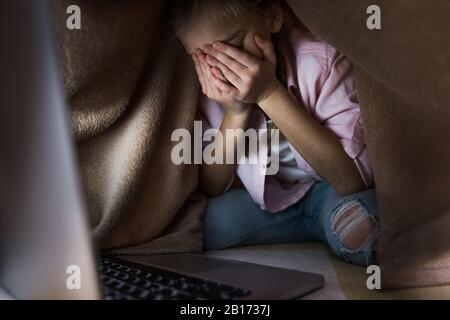 Selektive Fokussierung von frustrierten Kindern, die mit einer Decke bedeckt sind, während sie in der Nähe eines Laptops weinen, Cyberbullying Konzept Stockfoto